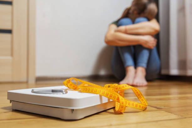 A woman sitting on the floor with her arms wrapped around her knees and a body weight scale and measuring tape sitting in front of her